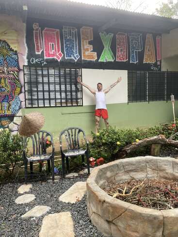 Um cara de camiseta regata branca e bermuda vermelha está de pé, com os braços estendidos, ao lado de um mural colorido, cadeiras de jardim, plantas e uma grande fogueira de pedra.