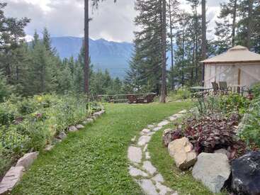 A scenic garden path winds through lush greenery, colorful flowers, large stones, and a round yurt, leading to wooden chairs overlooking forested mountains. Peaceful atmosphere.