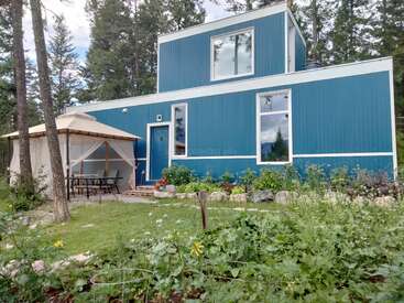 A blue modern house with white trim sits in a forested area. There's a gazebo with a table and chairs. A lush garden grows in front.