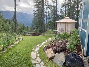A peaceful garden features a stone path, lush plants, a blue building, gazebo, forested mountain view, wooden chairs, and vibrant greenery under a cloudy sky.