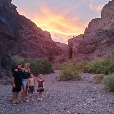 A family of four stands together in a rocky desert canyon at sunset, with orange clouds glowing above rugged cliffs and green bushes scattered around.