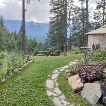 Un chemin de jardin pittoresque serpente à travers une verdure luxuriante, des fleurs colorées, de grosses pierres et une yourte ronde, menant à des chaises en bois surplombant des montagnes boisées. Atmosphère paisible.