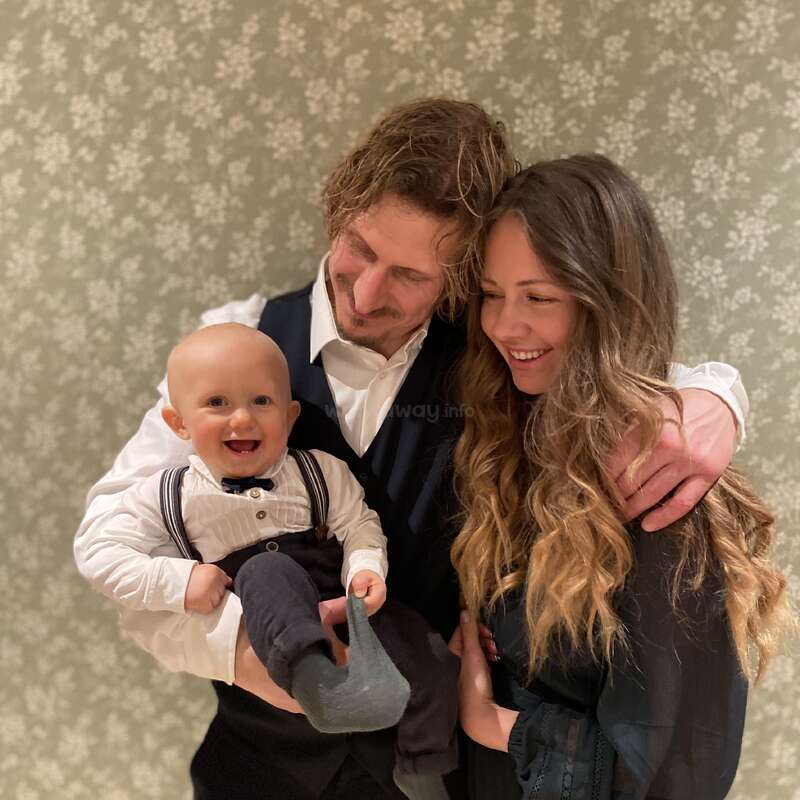 Uma família alegre está reunida, sorrindo calorosamente. O pai segura um bebê feliz vestido com suspensórios, enquanto a mãe o abraça com carinho em um fundo estampado.