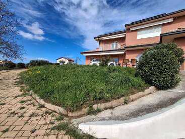 A modern peach-colored house with white shutters stands behind a grassy yard and paved driveway, under a vibrant blue sky with scattered clouds and lush greenery.