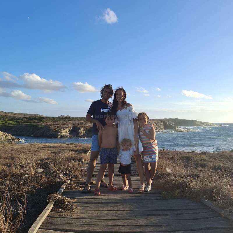 A family of five poses on a wooden path by the sea, smiling together under a bright blue sky with scattered clouds and beautiful coastal scenery.