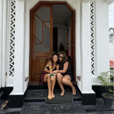 Two women are sitting barefoot on the front steps of a white building with wooden doors, smiling and enjoying each other's company on a relaxed day.