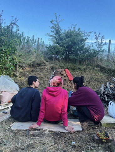 Tres personas están sentadas al aire libre sobre tablas de madera, de espaldas a la cámara, frente a una pequeña estructura de ladrillo, rodeadas de vegetación, aperitivos, madera y un cielo azul claro.