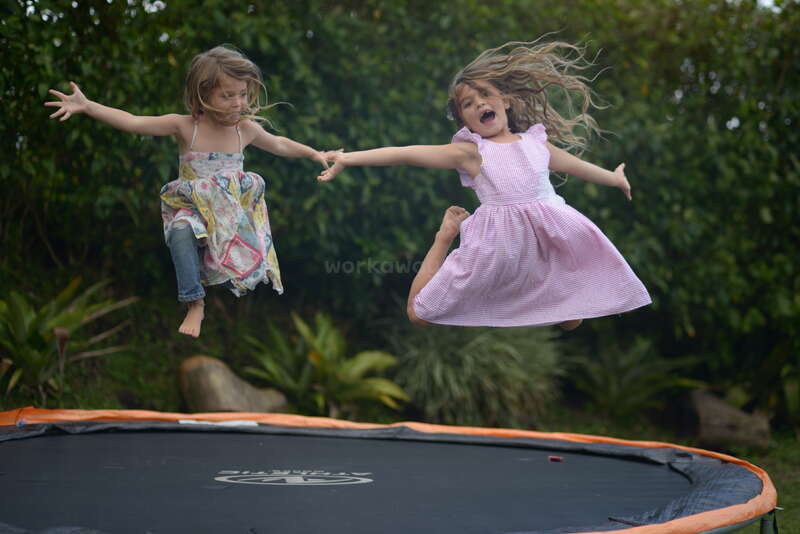 Zwei junge Mädchen mit langen Haaren springen auf einem Trampolin in einem Hinterhof, sie tragen Kleider und Jeans, mit einem unscharfen grünen Hintergrund.