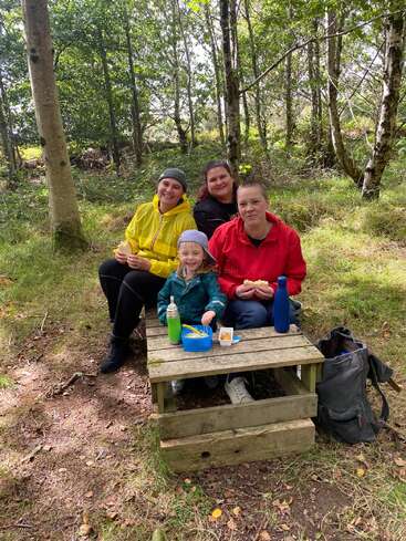Quatro pessoas sentam-se juntas em uma clareira na floresta, sorrindo e desfrutando de um piquenique. Em uma pequena mesa de madeira, você encontra lanches, bebidas e uma lancheira azul. Ambiente descontraído.