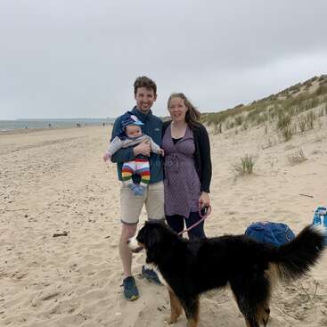 The image depicts a family of three, accompanied by their dog, standing on a sandy beach with a cloudy sky in the background. The family is dressed in casual attire.