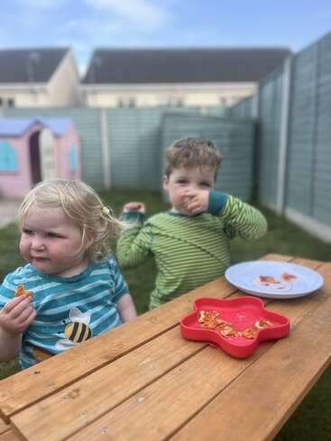 Two young children sit at a wooden picnic table outdoors, eating snacks. There are plates with food. Behind them is a fenced backyard and playhouse.