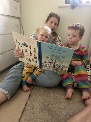 A woman sits on the floor, reading a book called "All Around Bustletown: Winter" to two young children in colorful pajamas, everyone looking engaged.