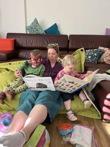A woman sits on the floor with two young children, reading books together. One child has their face painted like Spiderman. They look comfortable and cozy.