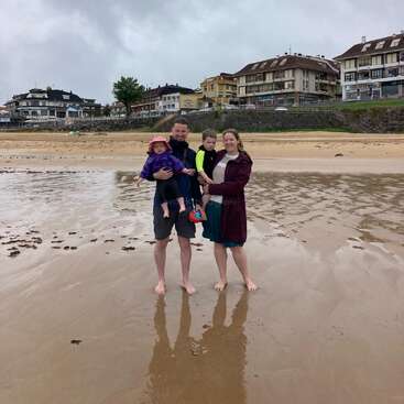 A family of four stands barefoot on a wet, sandy beach. They smile, wearing jackets. Colorful buildings and cloudy skies provide a calm, coastal backdrop.