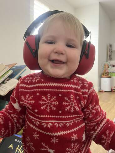 A smiling baby wearing red earmuffs and a festive red sweater with snowflake patterns stands indoors, surrounded by books and toys, radiating joy and cuteness.