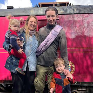 A happy family of four poses in front of a bright pink train. The adults smile while holding two young children, enjoying a fun outdoor day together.