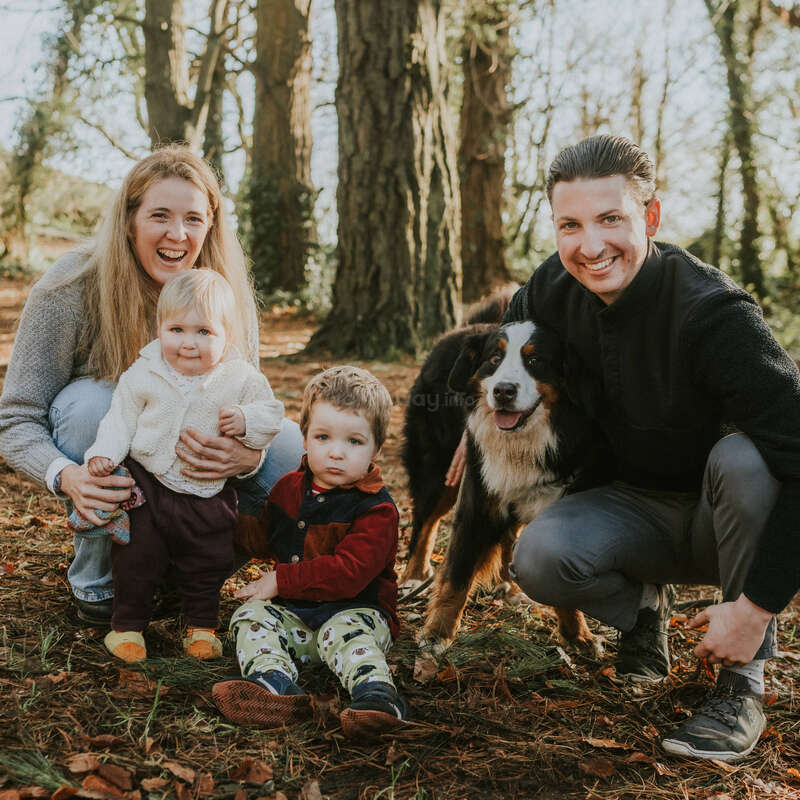 A happy family poses outdoors in a forest. The mother and father smile, their two young children and a large, friendly dog are gathered together.