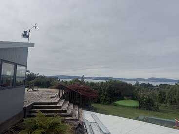A cloudy sky hovers over a lakeside scene with distant hills. In the foreground, there’s a deck, garden, and green landscape surrounding a modern house.
