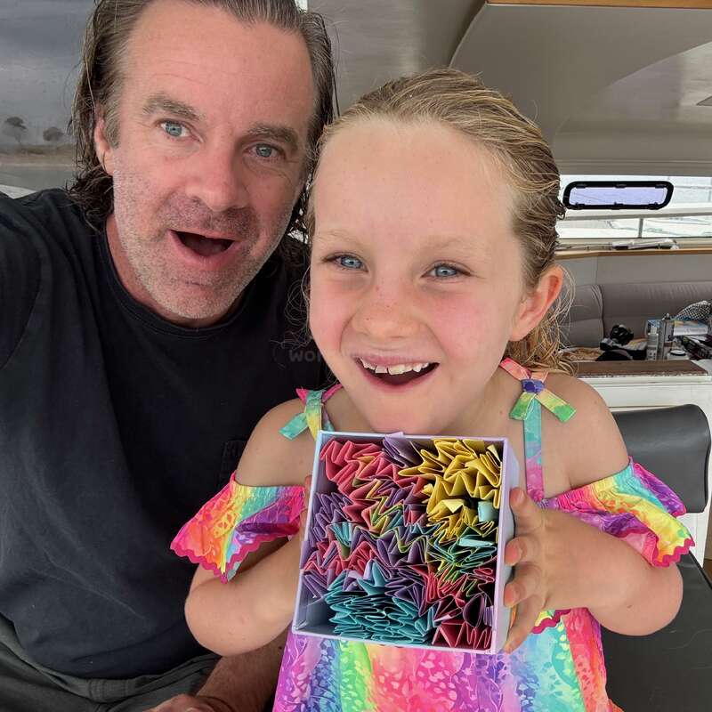 A man and a smiling young girl hold a box of colorful folded paper stars, enjoying a joyful moment together on what looks like a boat.