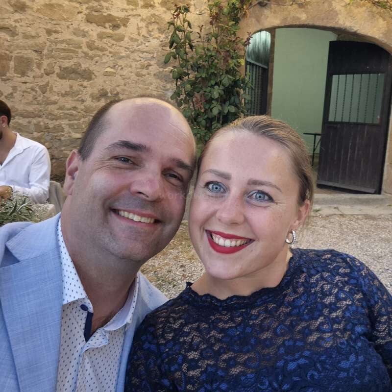 Un couple souriant prend un selfie en extérieur, devant un mur de pierre rustique. La femme porte un haut en dentelle bleu marine, tandis que l'homme porte une veste bleu clair.