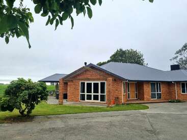 Das Bild zeigt ein einstöckiges rotes Backsteinhaus mit einem grauen Dach, einer großen Eingangstür, Fenstern und einer überdachten Veranda vor einem bewölkten Himmel.