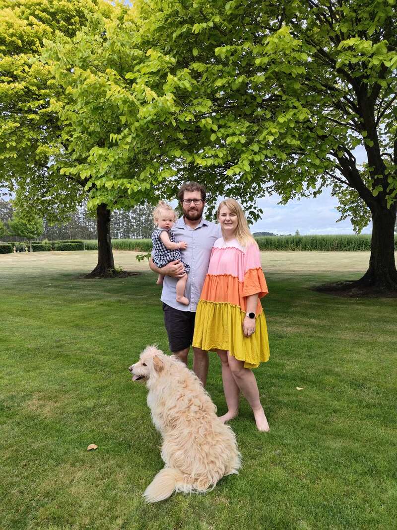 Das Bild zeigt eine dreiköpfige Familie, bestehend aus einem Mann, einer Frau und einem Baby, die mit einem Hund auf einer saftigen Wiese stehen, umgeben von Bäumen und einem Maisfeld im Hintergrund.