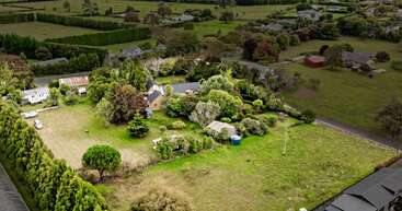 Uma propriedade rural cercada por vegetação, com uma casa, anexos, um amplo gramado, árvores maduras e jardins. As casas vizinhas e os campos abertos completam a paisagem tranquila.