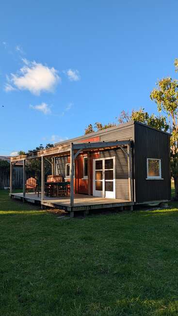 Uma pequena cabana moderna com um deck de madeira e uma área de estar ao ar livre, situada sobre um gramado verde, cercada por árvores, sob um céu azul claro com nuvens.