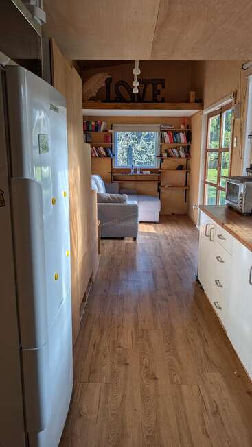 Esta aconchegante casinha tem piso de madeira, uma cozinha compacta, uma área de estar bem iluminada com um sofá cinza, estantes de livros e uma placa decorativa com a palavra “LOVE” pendurada acima.