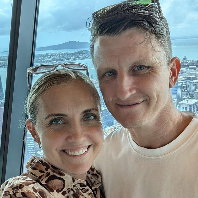 Um casal sorridente posa bem juntinho para uma selfie em frente a uma grande janela, com a paisagem urbana e o mar visíveis ao fundo, num dia nublado.