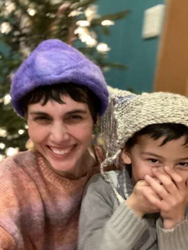 A woman and a child smile joyfully, wearing colorful hats. The child giggles, covering their mouth. A blurred Christmas tree with lights is in the background.