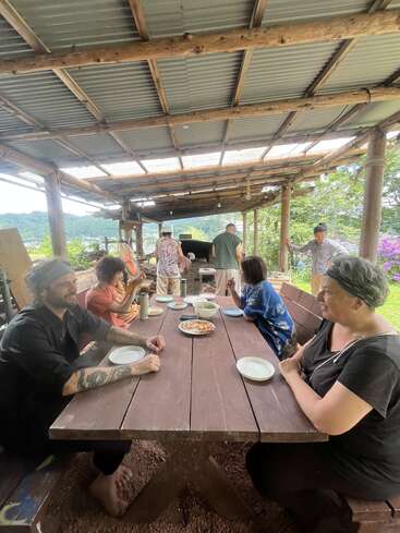 A group of people gather under a rustic wooden shelter, enjoying food and conversation at a long table, surrounded by greenery and a scenic outdoor setting.