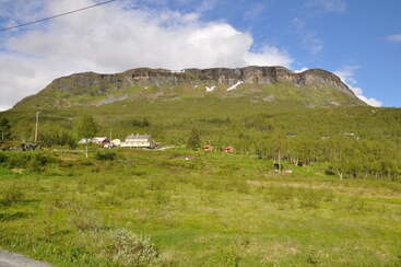 A imagem retrata uma paisagem serena com uma cadeia de montanhas com um pico proeminente, cercada por um corpo de água e uma área gramada, evocando uma sensação de beleza natural e tranquilidade.