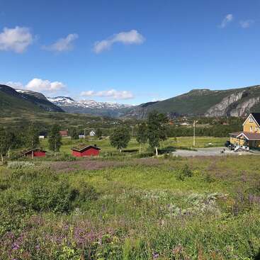 The image depicts a serene mountainous landscape with a vibrant field of wildflowers, featuring small houses and trees, set against a backdrop of majestic mountains under a clear blue sky.