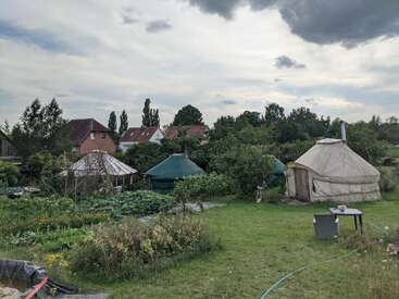 A imagem mostra vários yurts em um jardim verde e exuberante. Há hortas, casas ao fundo e um céu parcialmente nublado acima de você.