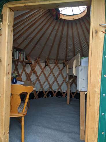 Este é o interior de uma yurt aconchegante com vigas de madeira, uma claraboia, uma mesa, cadeira, estantes, cama e piso acarpetado. Simples e convidativo.