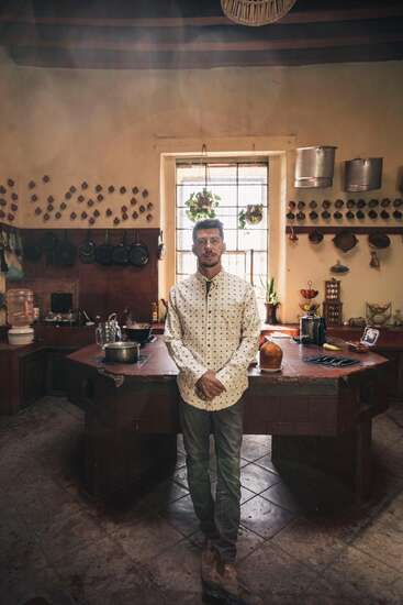 Un hombre se encuentra en una cocina rústica con macetas colgantes, luz solar que entra por una ventana, plantas, utensilios y alimentos ordenados en estantes y mostradores.