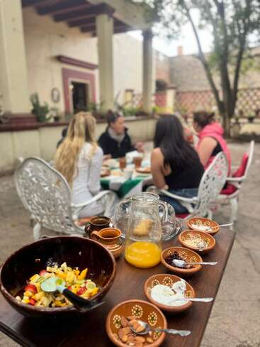 Un grupo de personas disfruta de una comida al aire libre en un encantador patio. La mesa muestra ensalada de frutas, zumo, frutos secos, yogur y semillas en cuencos de barro rústico.