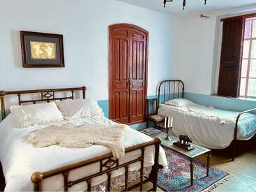 Un acogedor dormitorio con dos camas de estructura metálica, ropa de cama blanca, un armario de madera, obras de arte enmarcadas, una ventana, una alfombra vintage y una mesita de café de cristal.