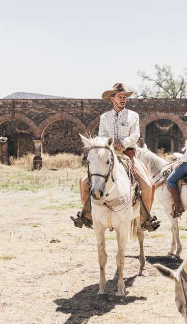 A man in a patterned shirt and cowboy hat sits on a white horse outdoors, with old stone arches and dry grass in the background, under a clear sky.