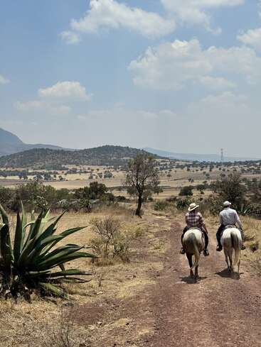 Two people ride horses along a dusty trail through dry, open countryside under a partly cloudy sky, surrounded by agave plants, trees, and distant hills.