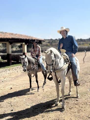 Two people wearing cowboy hats and casual western attire ride horses outdoors on a sunny day, enjoying a rustic ranch setting with clear skies and relaxed smiles.
