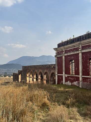 An old, weathered building with red walls and arches stands in a grassy field. Mountains rise in the distance under a partly cloudy sky. Serene atmosphere.