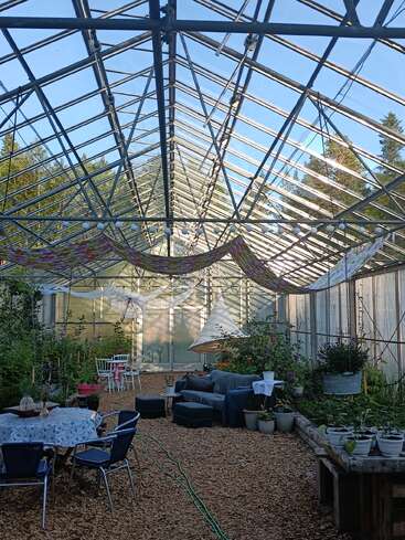 This bright greenhouse features a transparent roof, garden beds, hanging plants, cozy seating areas, tables, chairs, festive fabric decorations, and string lights, creating a welcoming atmosphere.