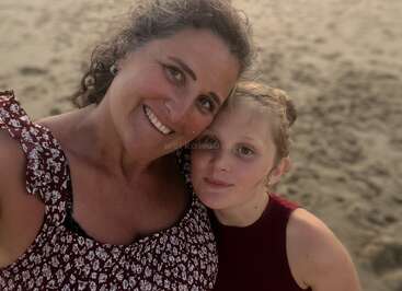 A woman and a young girl smile for a close-up selfie on a sandy beach, capturing a warm, loving moment together in natural sunlight.