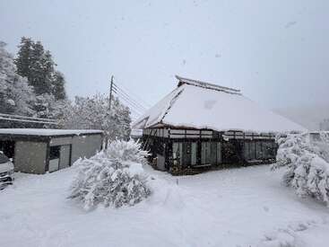 A traditional house with a steep thatched roof is blanketed in thick snow. Snow-covered trees and bushes surround the peaceful winter scene, snowflakes falling.