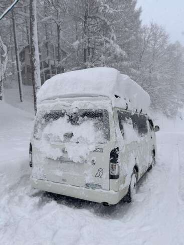 A van is completely covered in thick snow, parked on a snowy road. Snow continues to fall heavily, and the surrounding pine trees are frosted white.
