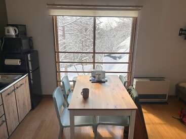 A cozy dining area with a wooden table, four chairs, and a mug. Large window shows snowy trees outside. Kitchen appliances and wooden floor are visible.