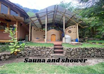 A rustic outdoor sauna and shower structure with a stone path and steps, surrounded by lush greenery, bamboo posts, and natural landscaping. Text reads, “Sauna and shower.”