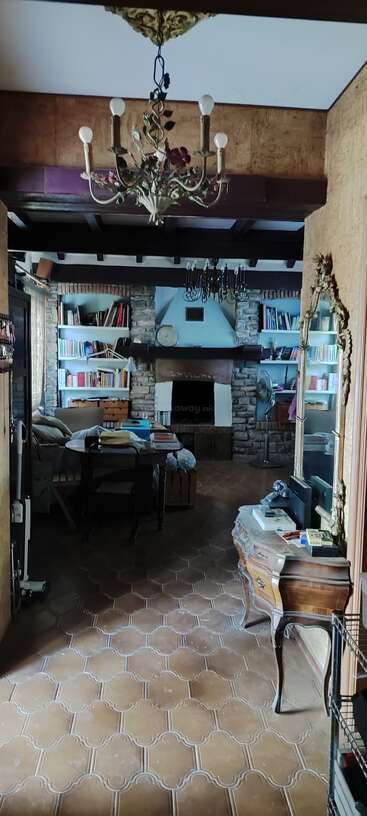 Das ist ein gemütliches, rustikales Wohnzimmer mit einem Steinkamin, Holzbalken, Bücherregalen, Vintage-Möbeln, Kronleuchtern, einem dekorativen Spiegel und einem warmen Fliesenboden. Eine einladende und heimelige Atmosphäre.